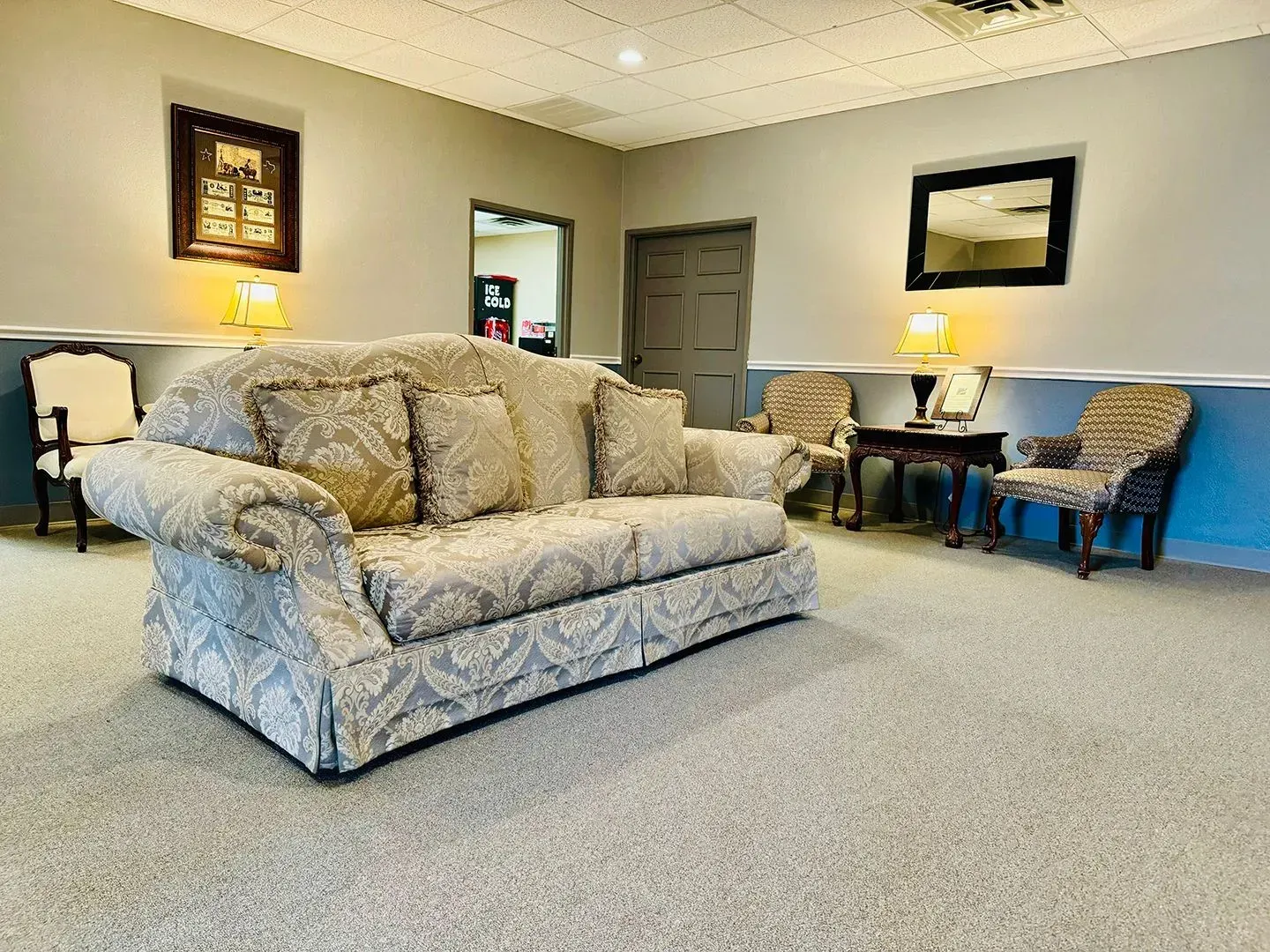 A waiting room with a patterned sofa, two armchairs, a side table with a lamp, a wall mirror, and a framed sign.