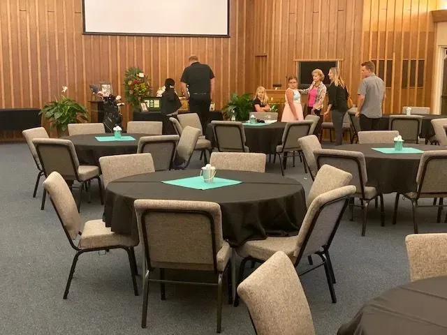 People gather in a room with wood-paneled walls, featuring round tables with black tablecloths and teal accents.