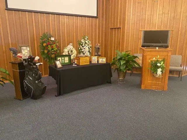 Memorial display with golf clubs, flowers, framed photos, and an urn on a table, beside a wooden podium in a room.
