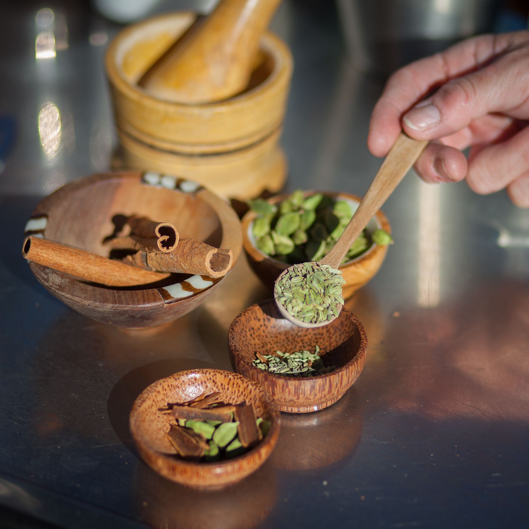Person scooping herbs from a wooden bowl with a small spoon, surrounded by other spices.