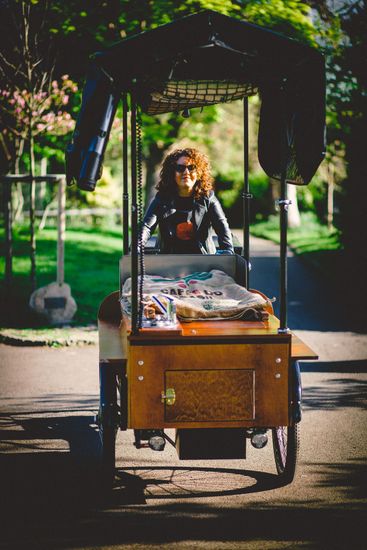 Woman with curly red hair operates a wooden vending cart on a path in a park, with a black awning overhead.