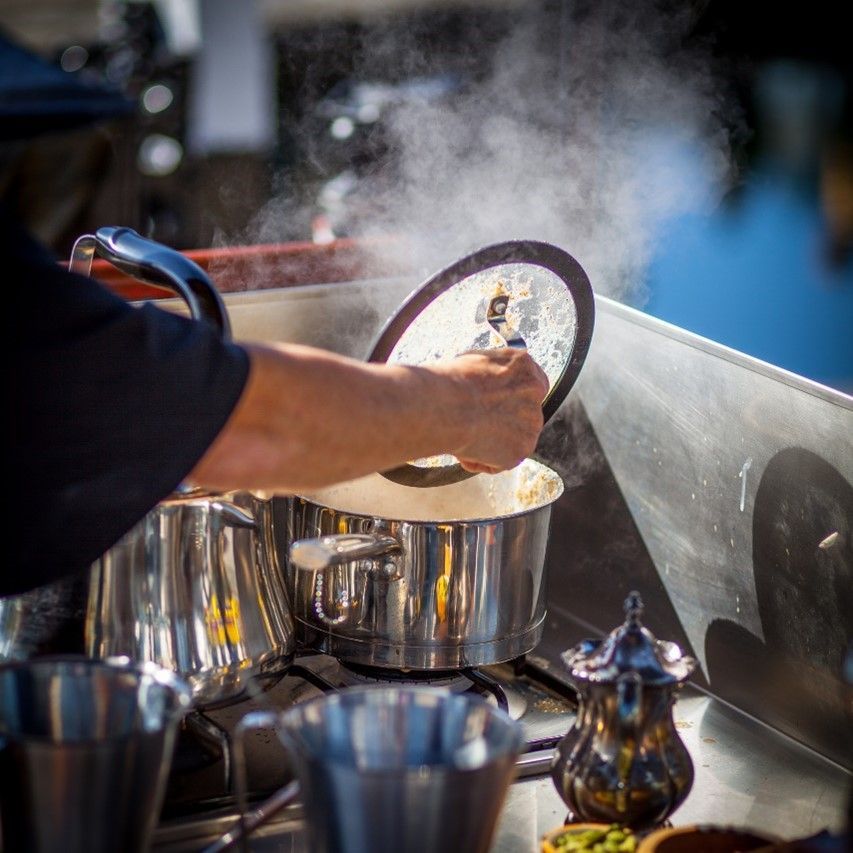 Person lifts lid from steaming pot on a stove, steam rising; outdoor market.