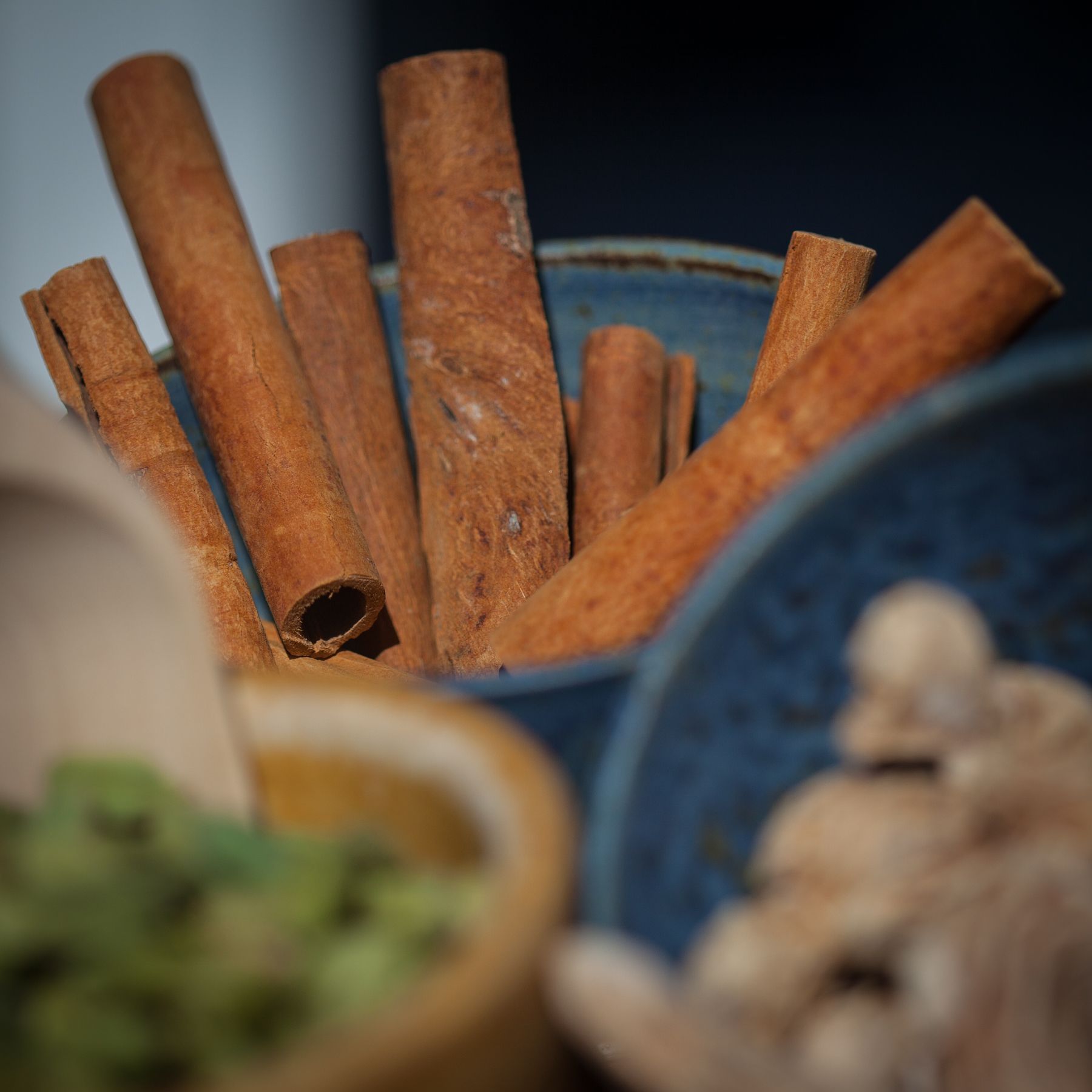 Cinnamon sticks in a blue ceramic bowl, with other spices in blurred containers in the foreground.