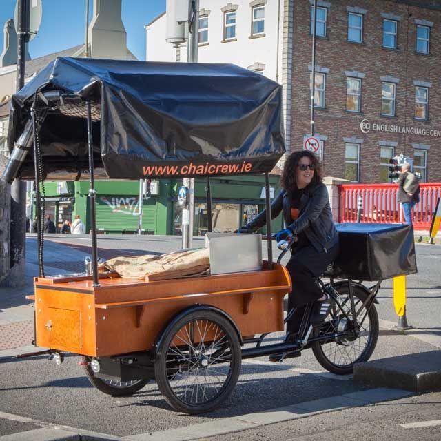 Woman riding a Chai Crew tricycle cart on a city street. Brown cart, black canopy, building in the background.