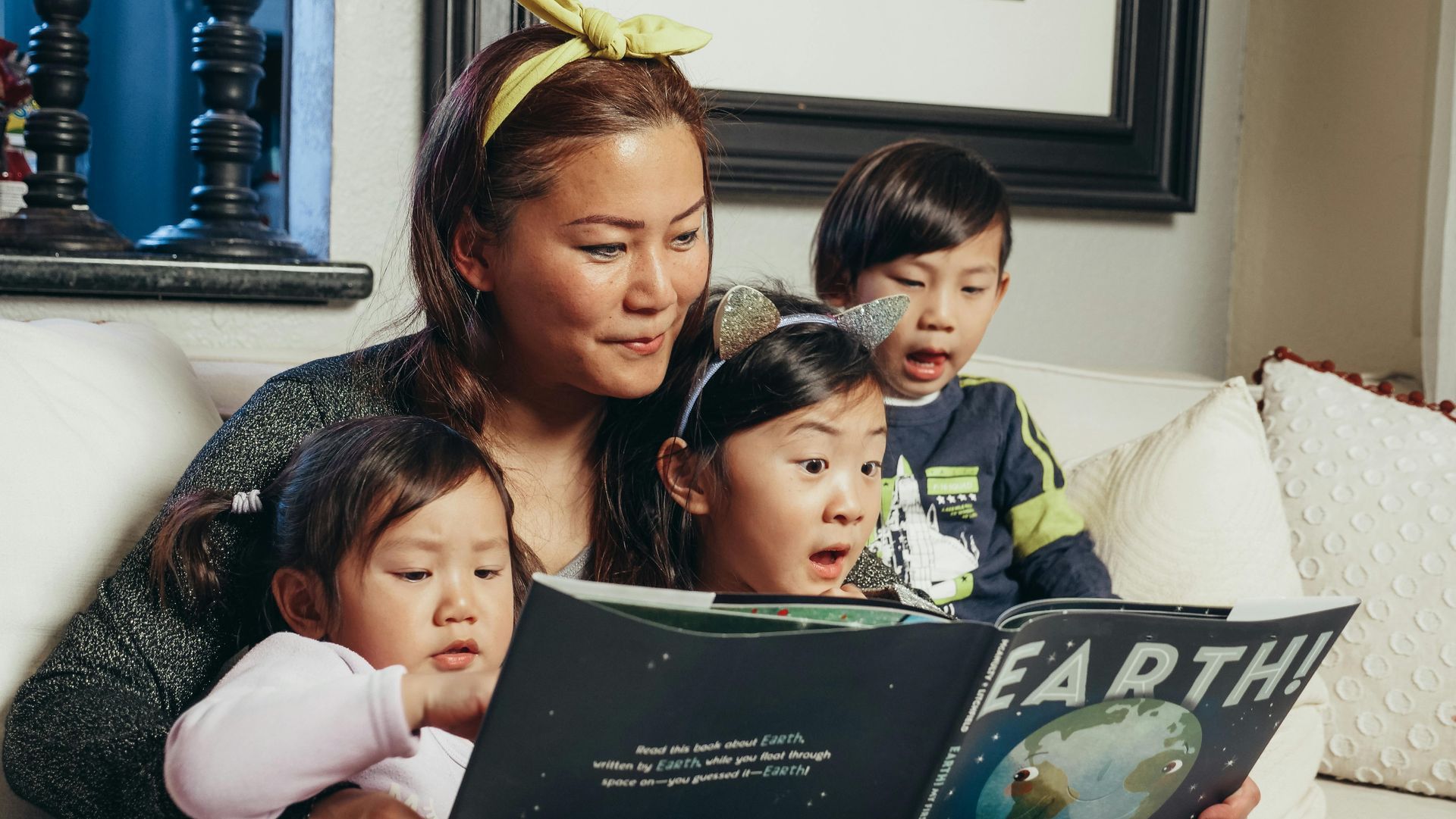 A woman is reading a book to three children while sitting on a couch.