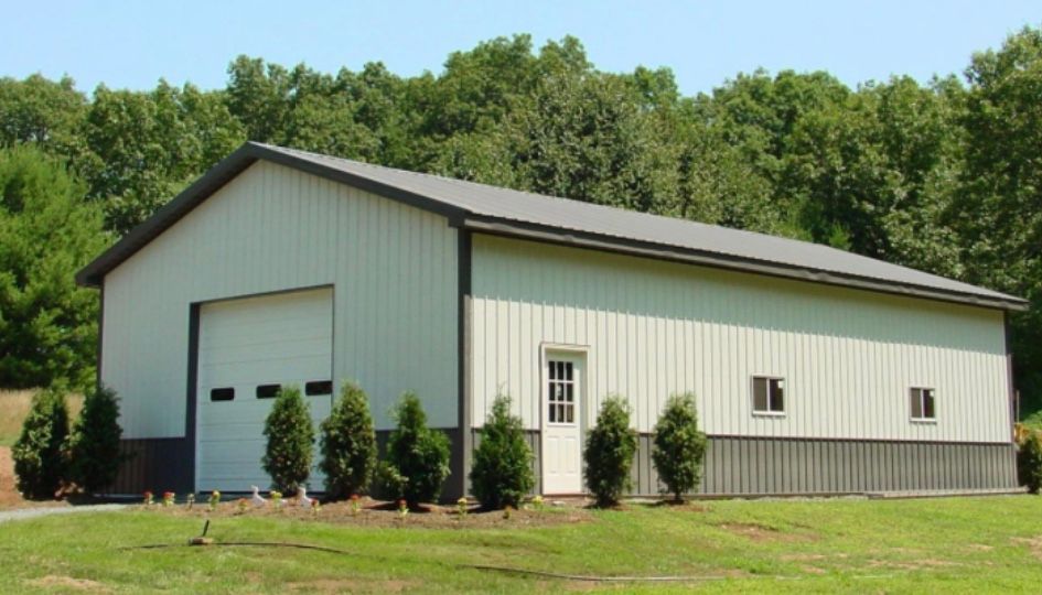 A large, white metal building with a garage door and small windows, set in a grassy yard against a backdrop of trees.