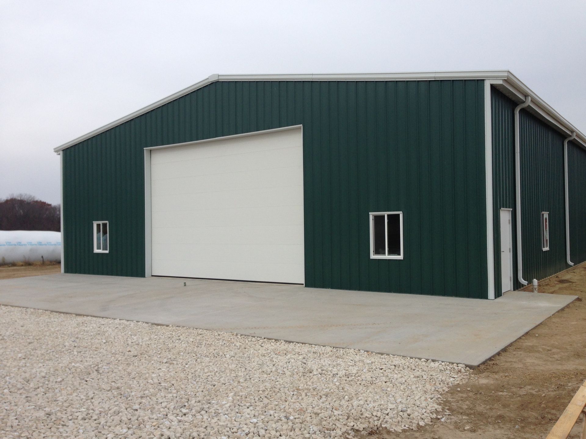 Green metal warehouse building with white garage door, concrete pad, and gravel.