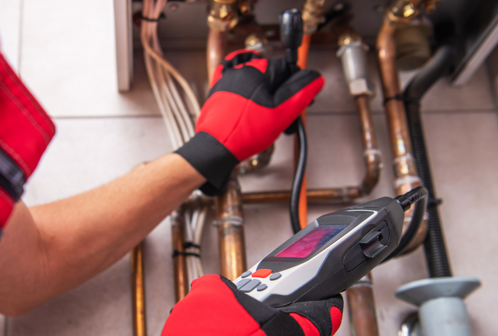 A person wearing red gloves inspects plumbing with a handheld device near a wall-mounted boiler.