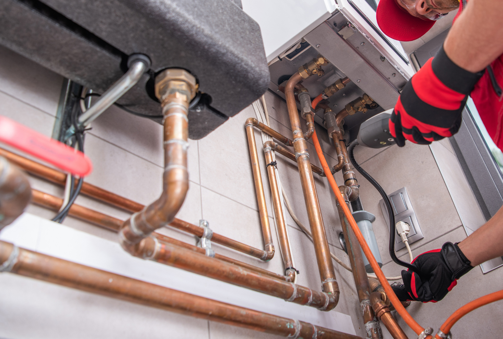 Plumber working on copper pipes and a boiler, wearing gloves and a red cap, indoors.