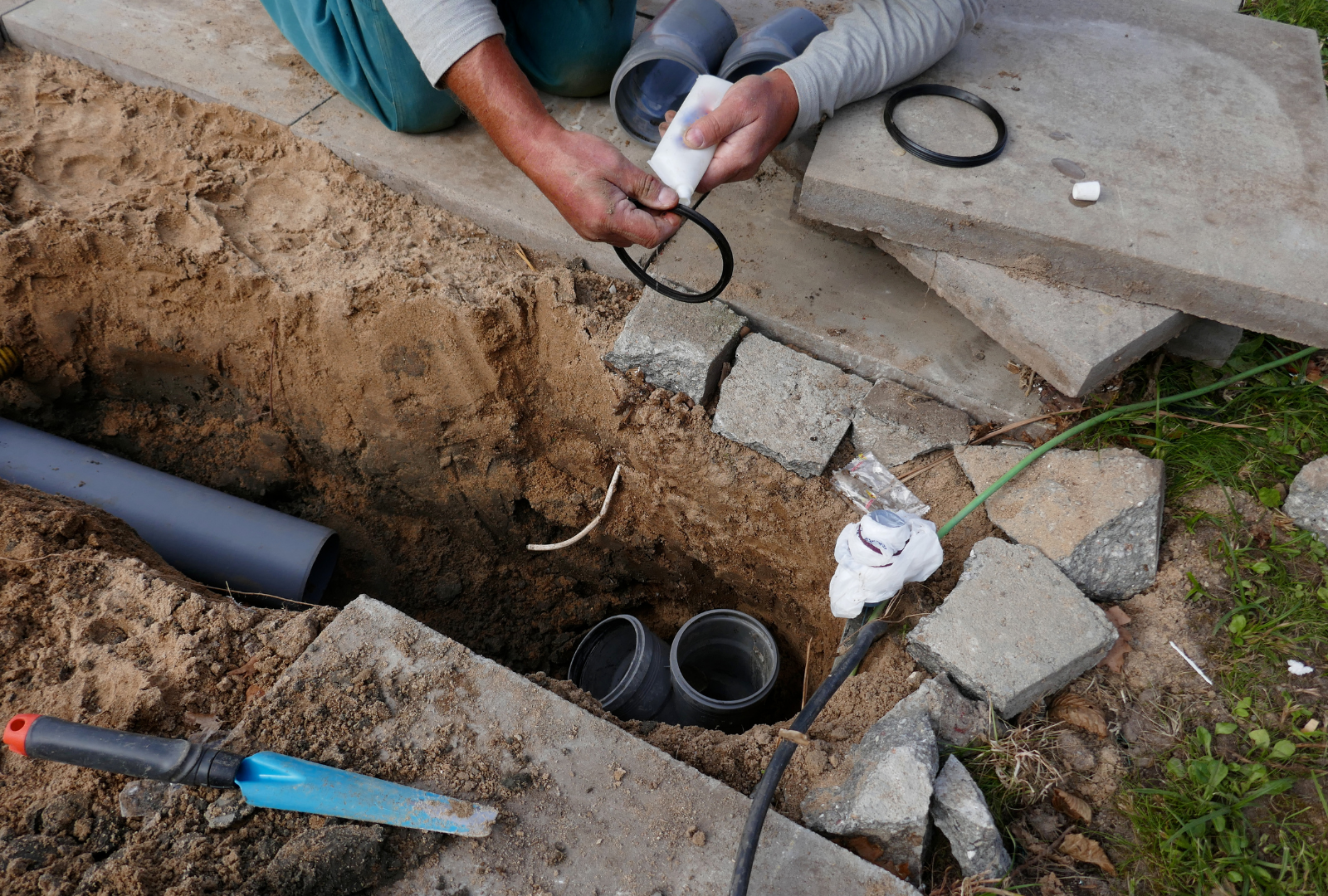 Man installing plumbing in a trench, holding an O-ring. Pipes, dirt, and concrete blocks are present.