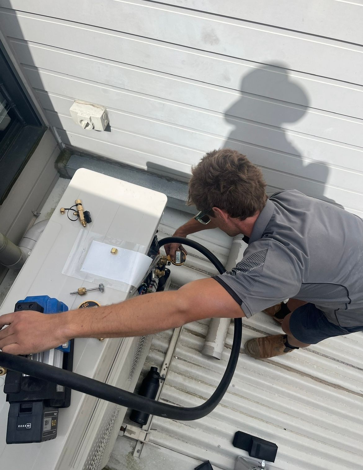A technician in a gray uniform works on an outdoor HVAC unit on a corrugated metal roof — BTM Electrical & Air in Burleigh Waters, QLD