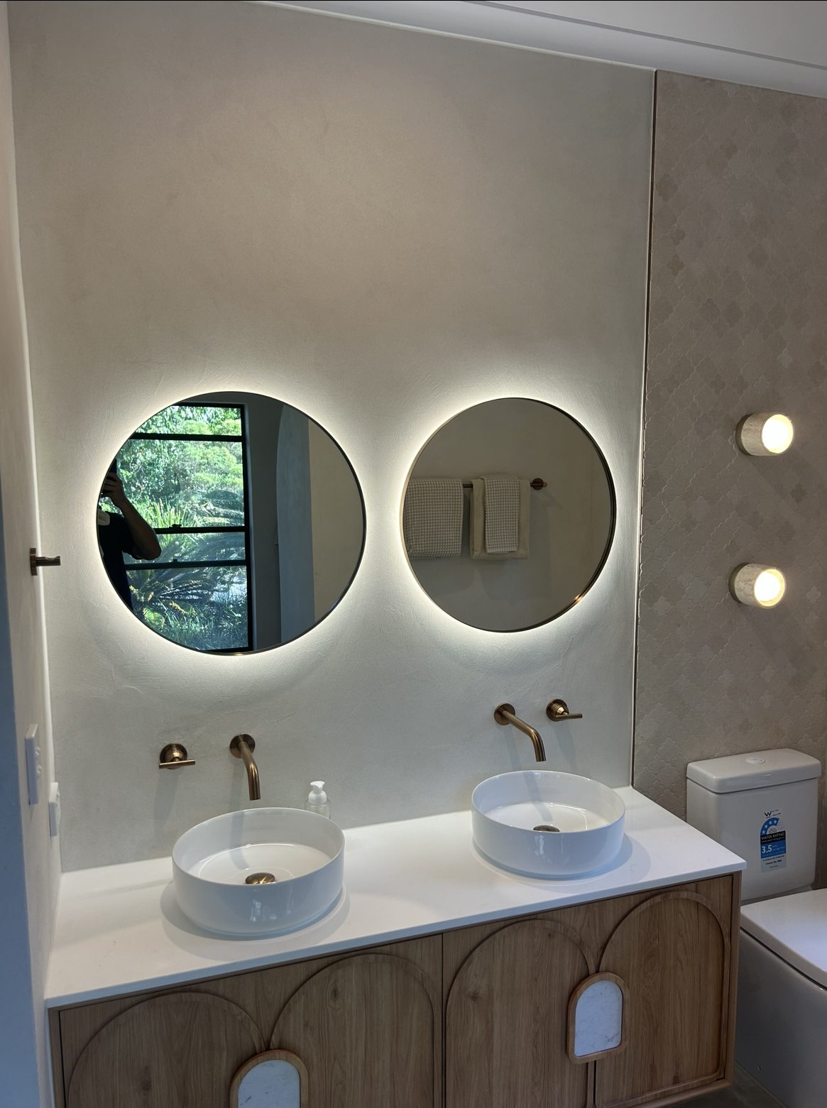 A modern double-sink bathroom vanity with round illuminated mirrors, brass fixtures, and a wood-arched cabinet. — BTM Electrical & Air in Burleigh Heads, QLD