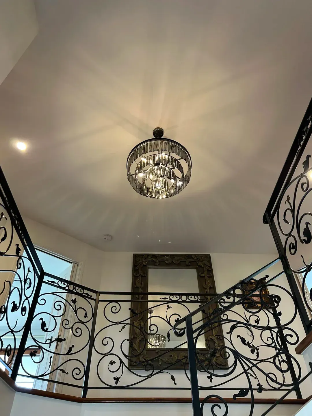 An ornate, black wrought-iron staircase railing in a foyer with a crystal chandelier and a large, framed wall mirror. — BTM Electrical & Air in Currumbin, QLD