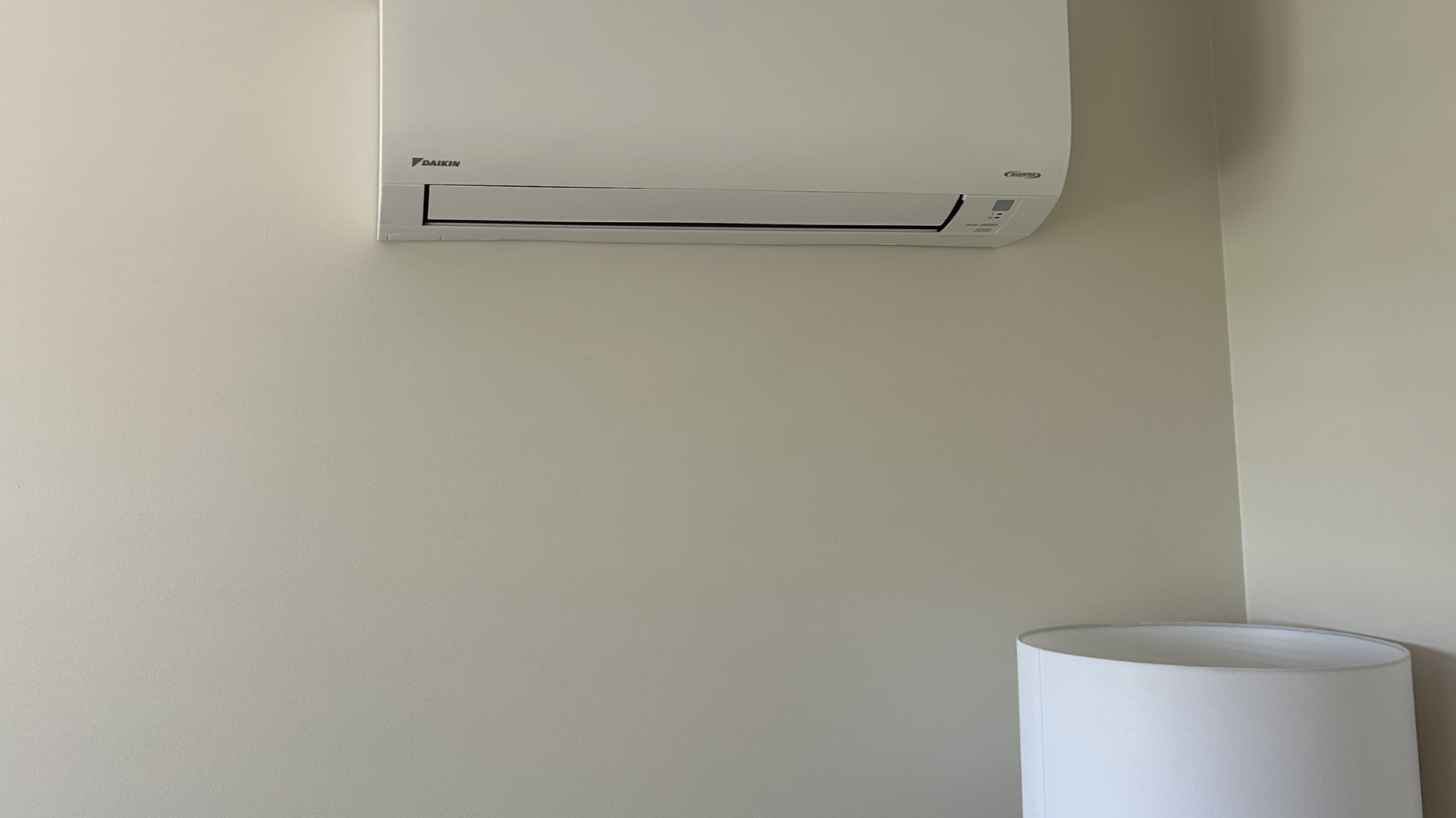 A white wall-mounted air conditioning unit installed high on a plain, beige wall, with a white lampshade in the corner. — BTM Electrical & Air in Coolangatta, QLD