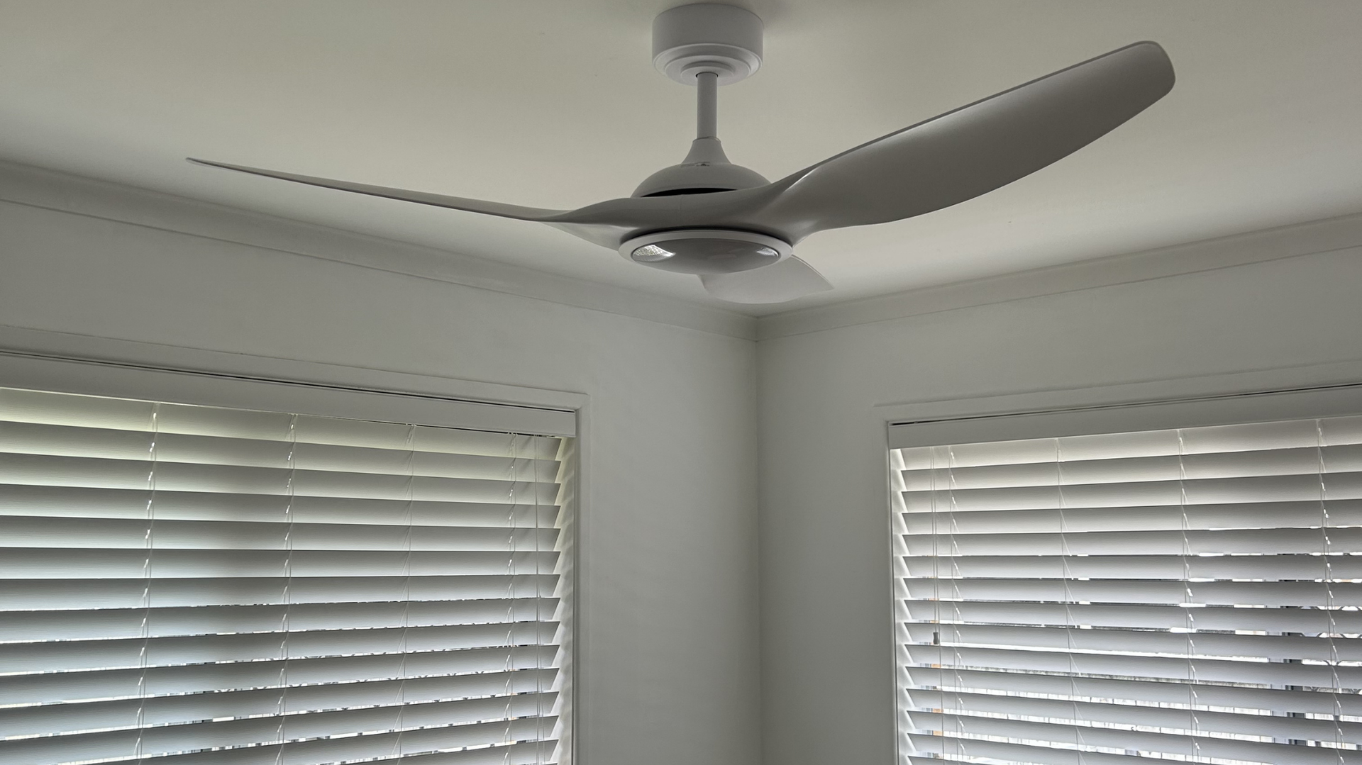 A white three-blade ceiling fan is centered in a room with white walls and windows covered by white horizontal blinds. — BTM Electrical & Air in Southport, QLD