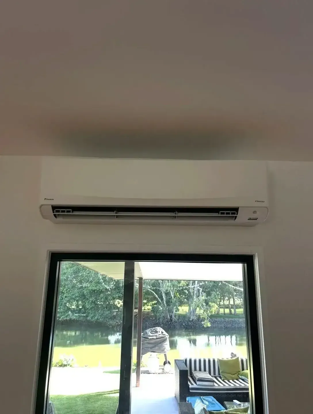 A white wall-mounted air conditioning unit installed above a window overlooking a patio and greenery.  — BTM Electrical & Air in Elanora, QLD