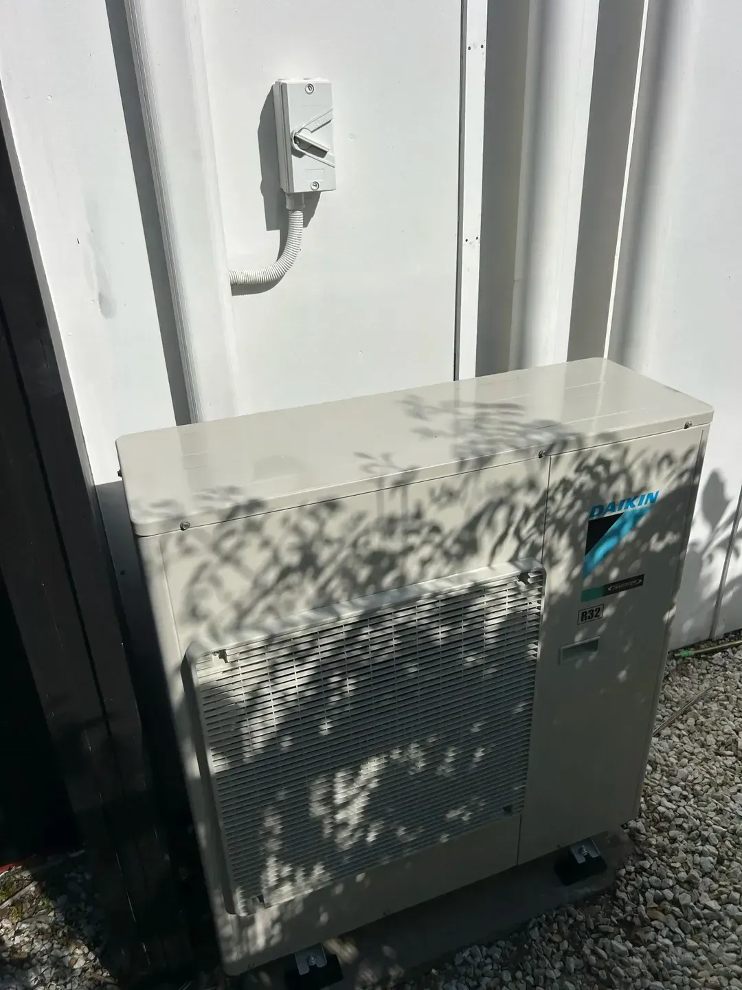 A beige outdoor air conditioning unit sits on gravel against a white wall with a mounted electrical disconnect switch. — BTM Electrical & Air in Burleigh Waters, QLD