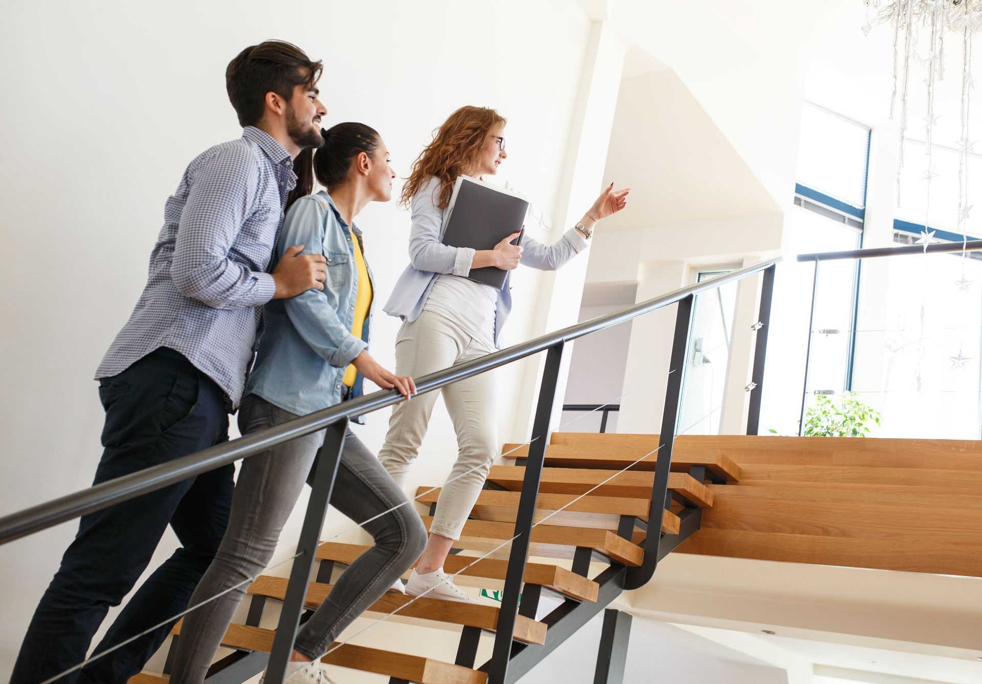 A real estate agent leads a couple up a wooden staircase, pointing toward a window.