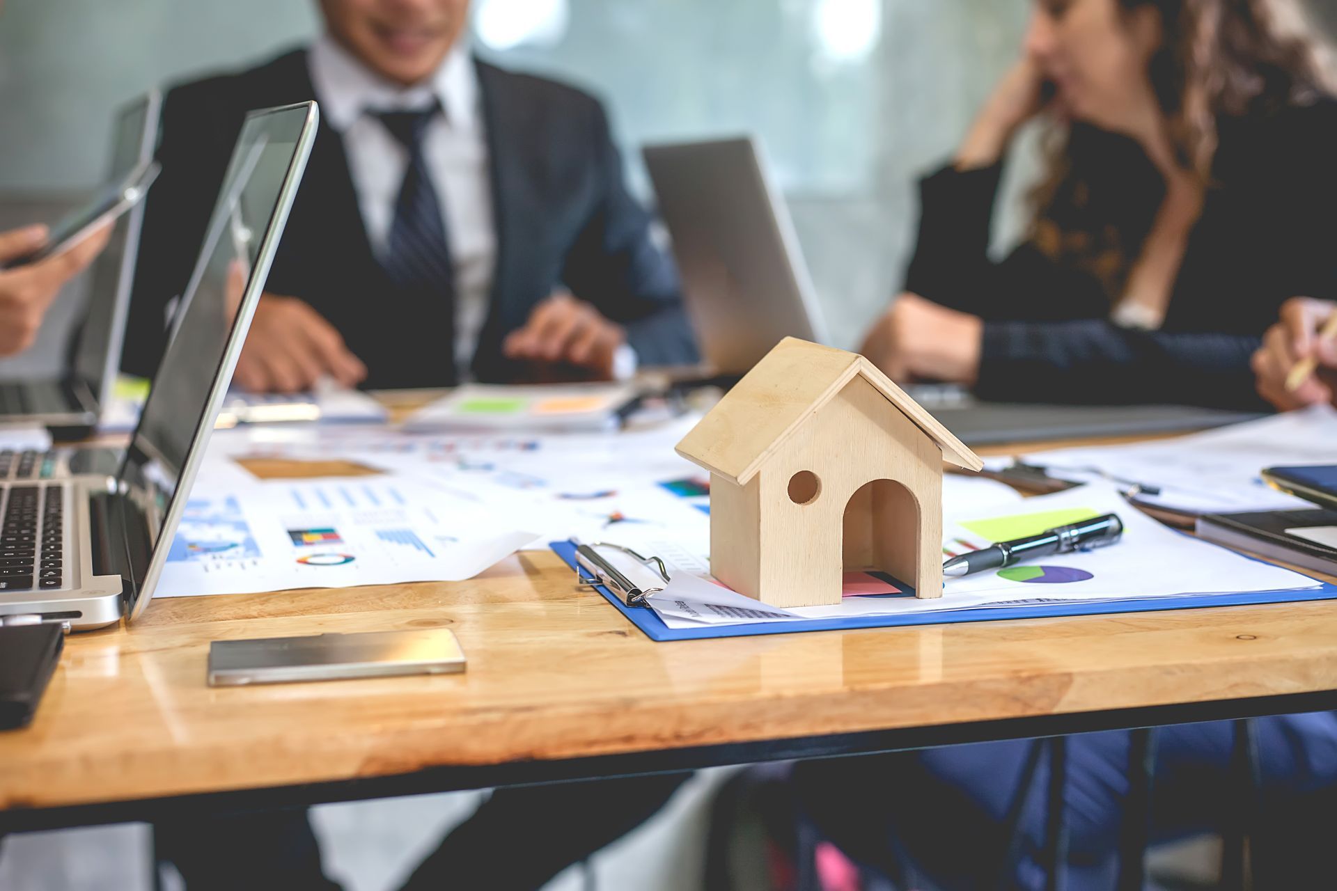 Wooden house model on a table with people, charts, and laptops, likely a real estate meeting.