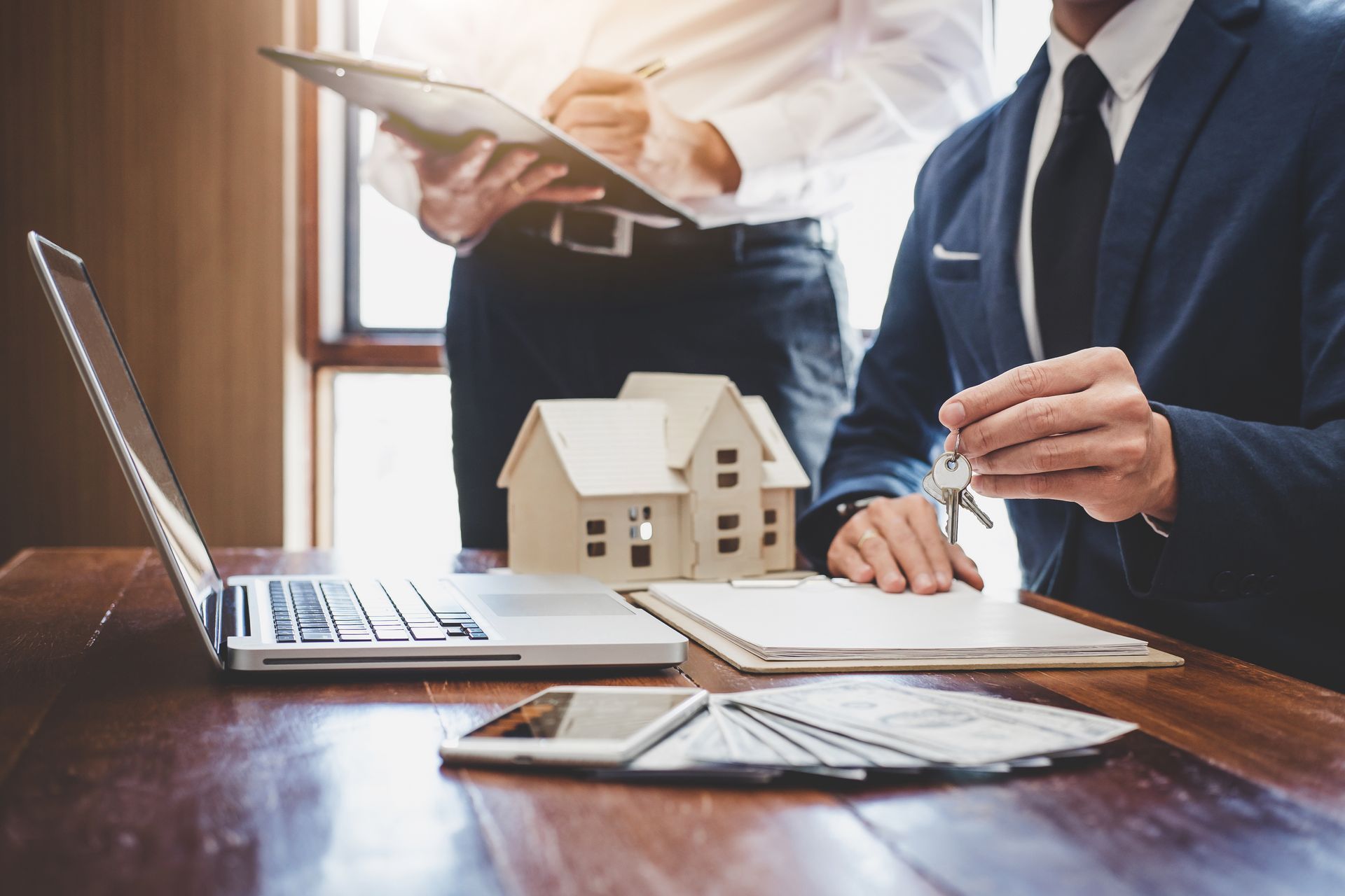Real estate agents at a table with a laptop, model house, money, and paperwork. One holds keys. Real estate agents at a table with a laptop, model house, money, and paperwork. One holds keys.
