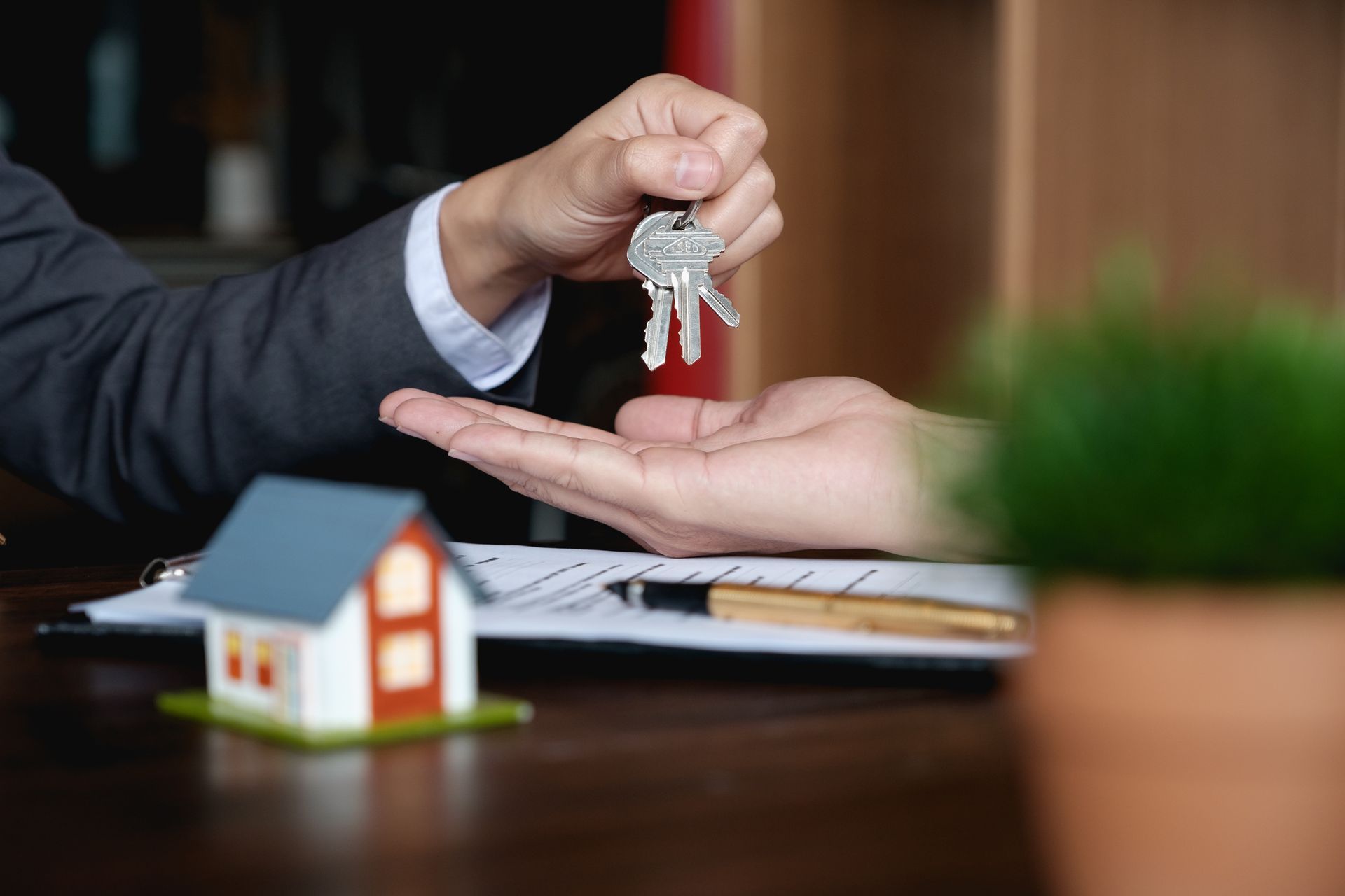 Person handing keys to another, symbolizing a real estate transaction. A house model and paperwork are on the table. Person handing keys to another, symbolizing a real estate transaction. A house model and paperwork are on the table.