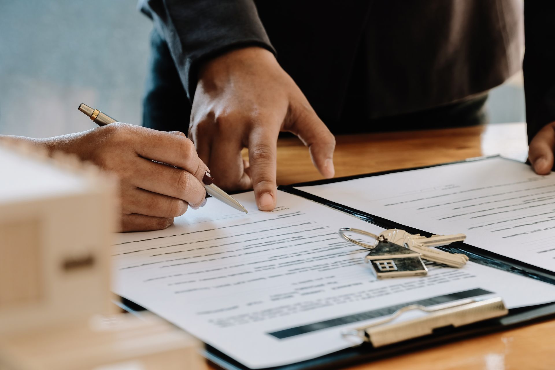 Hands signing a document, keys and a miniature house on a desk.