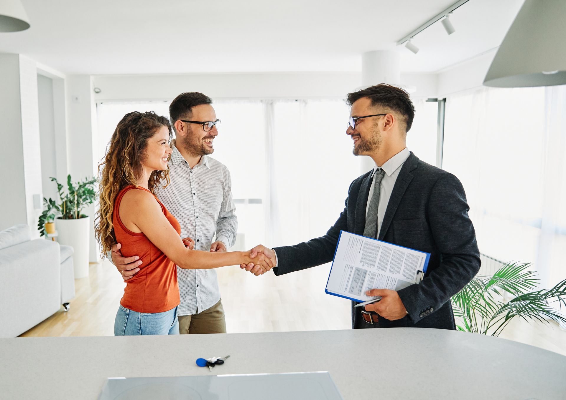Couple shaking hands with a realtor in a bright apartment; contract and keys on table.
