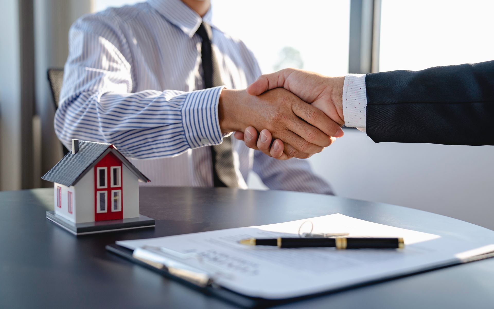 Two people shaking hands over a house model and real estate documents.