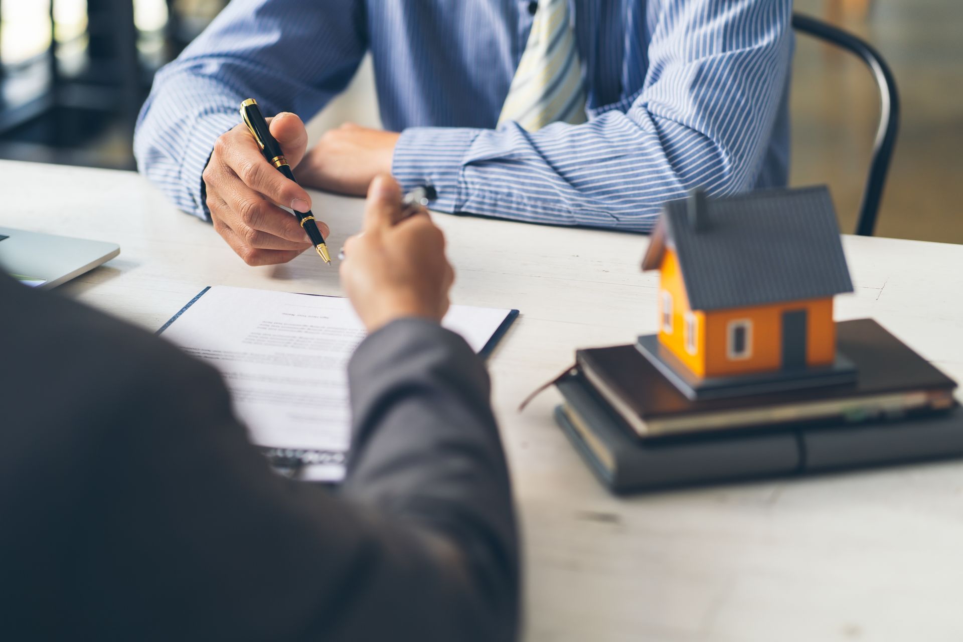 Two people at a table; one signing documents, the other gesturing. A small house model sits on books nearby. Two people at a table; one signing documents, the other gesturing. A small house model sits on books nearby.