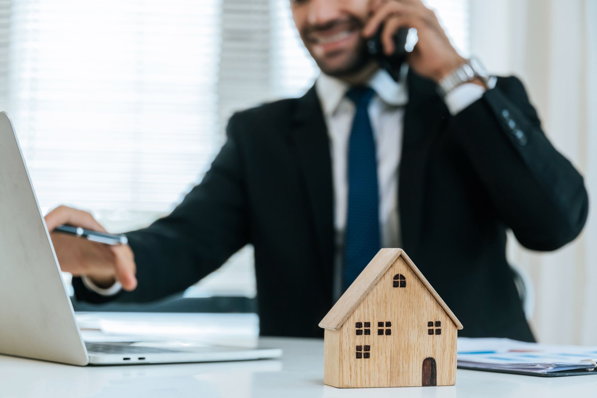 Real estate agent on phone, working on laptop with small wooden house on desk.
