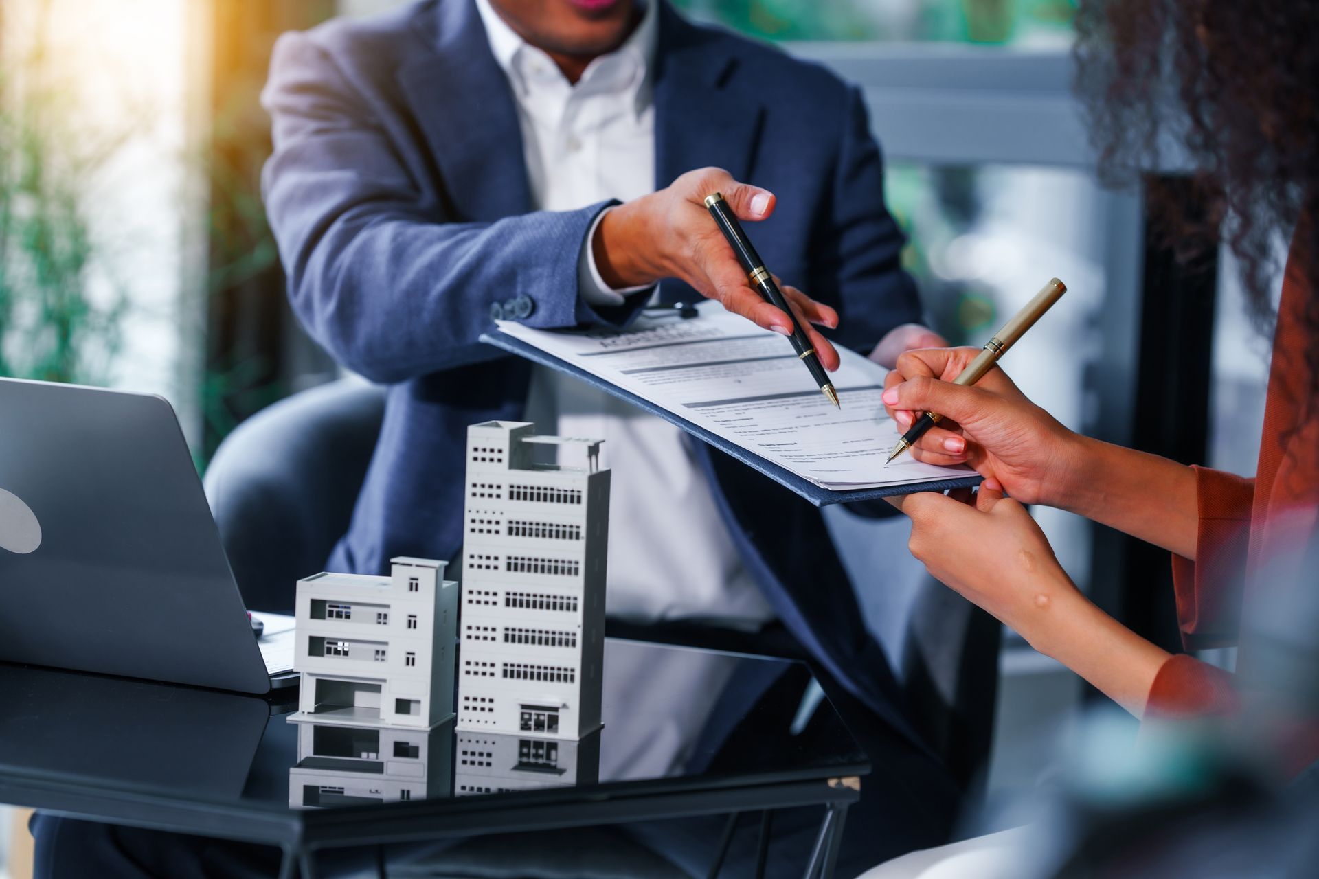 Real estate agent pointing at a document while a person signs. Building model on table. Real estate agent pointing at a document while a person signs. Building model on table.