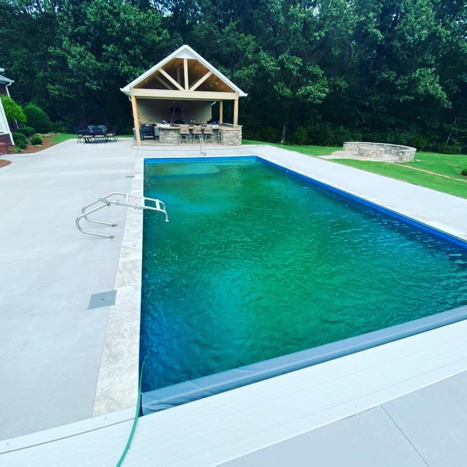 A rectangular pool with turquoise water, adjacent to a concrete patio and a wooden gazebo, surrounded by green grass and trees.