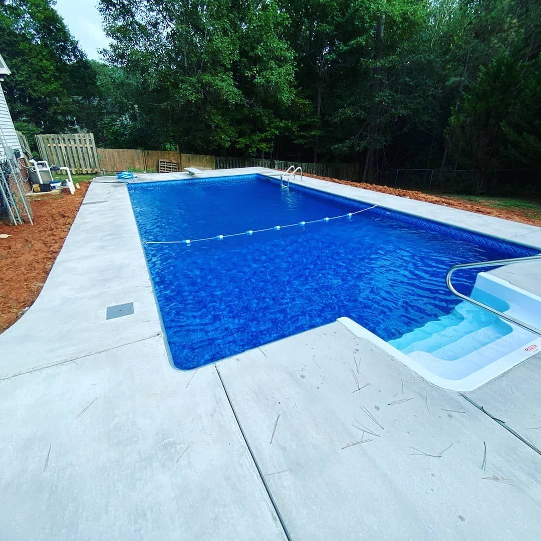 A rectangular pool with blue water, surrounded by concrete and trees. Steps lead into the shallow end.