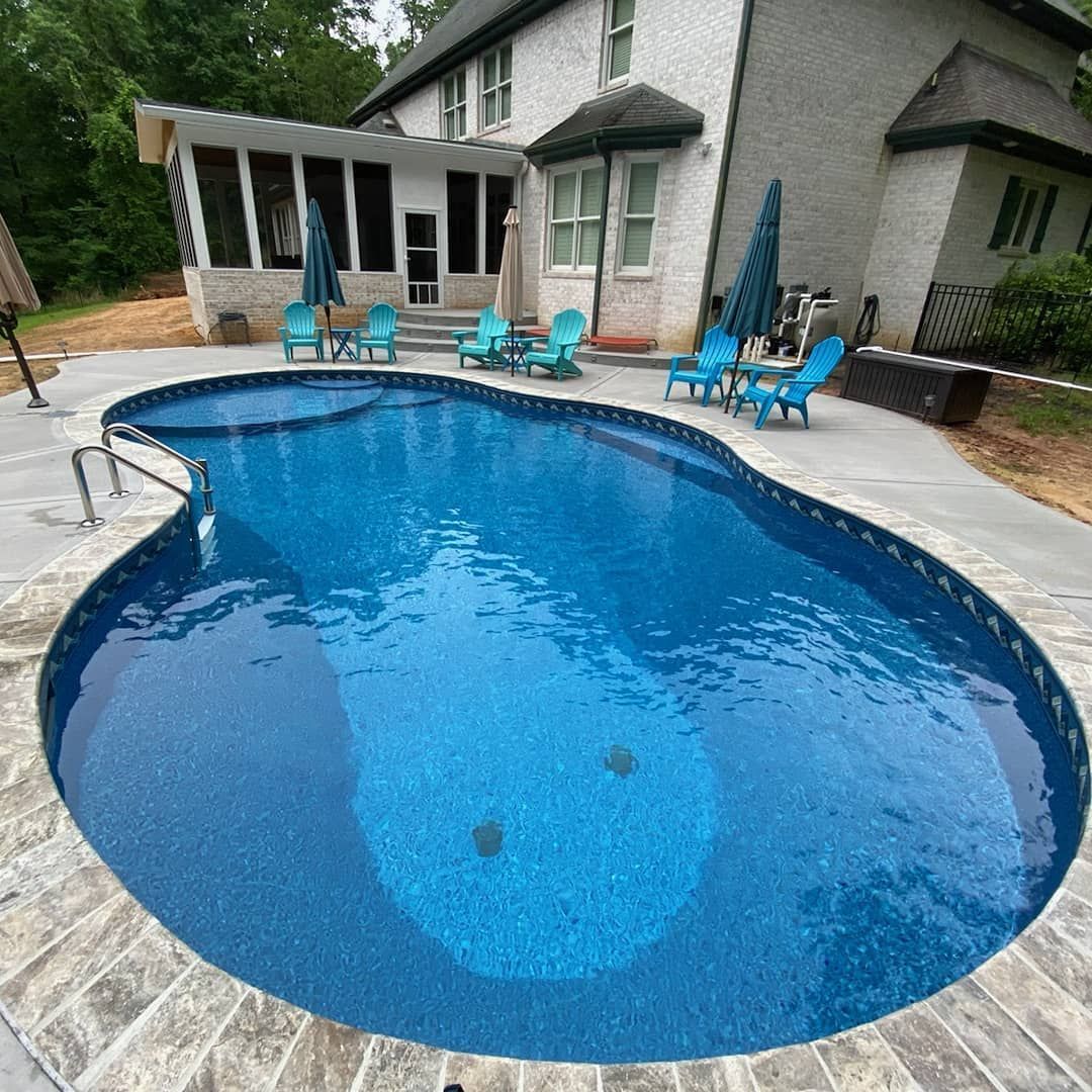 Swimming pool with blue water, surrounded by concrete patio and lounge chairs in backyard of a house.