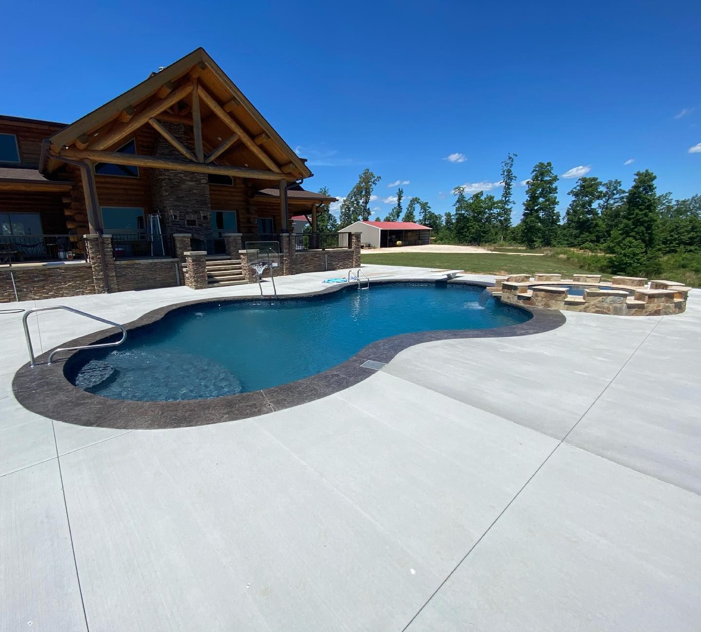Pool in front of log cabin home with blue sky, concrete deck, and a stone firepit.