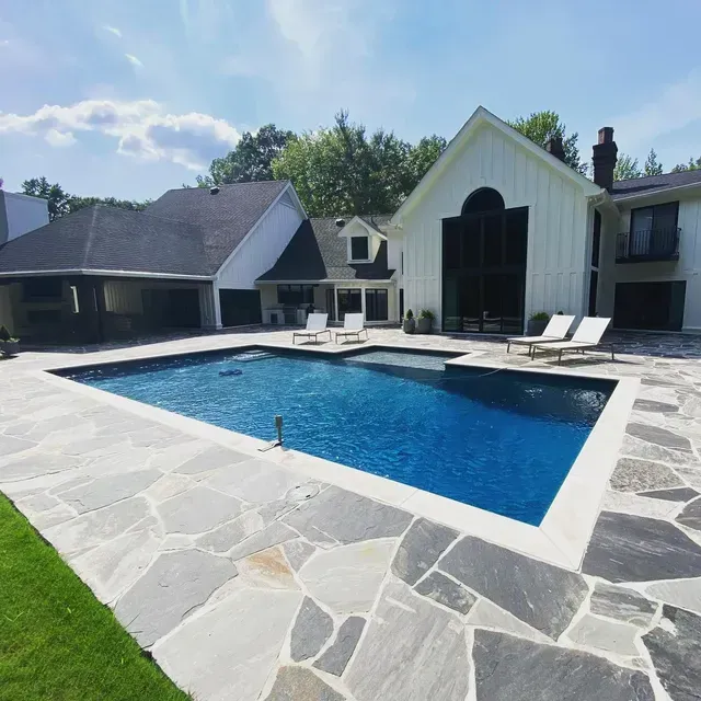 Backyard pool with flagstone patio, two lounge chairs, and white house with dark roof.