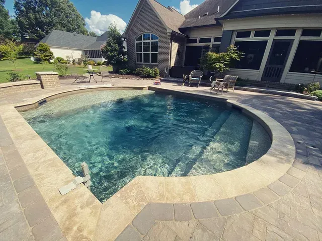 A backyard pool with light-colored stone edging, turquoise water, and a brick house in the background.