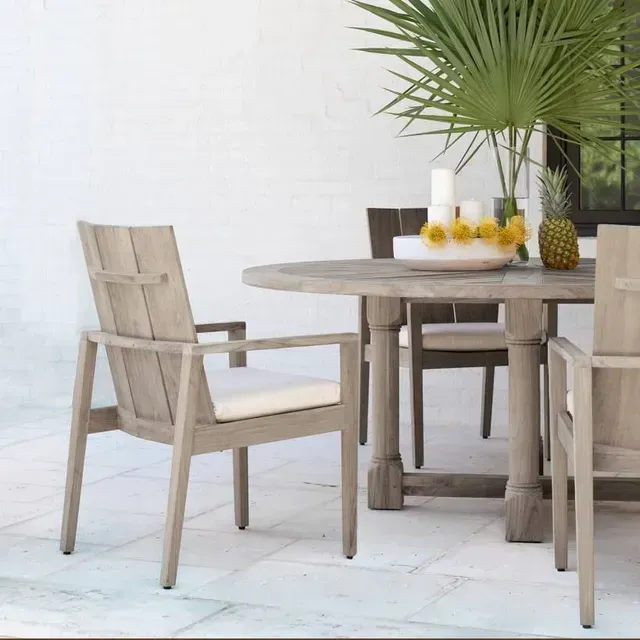 Outdoor dining set: round weathered wood table, chairs with cushions, potted palm, pineapple.
