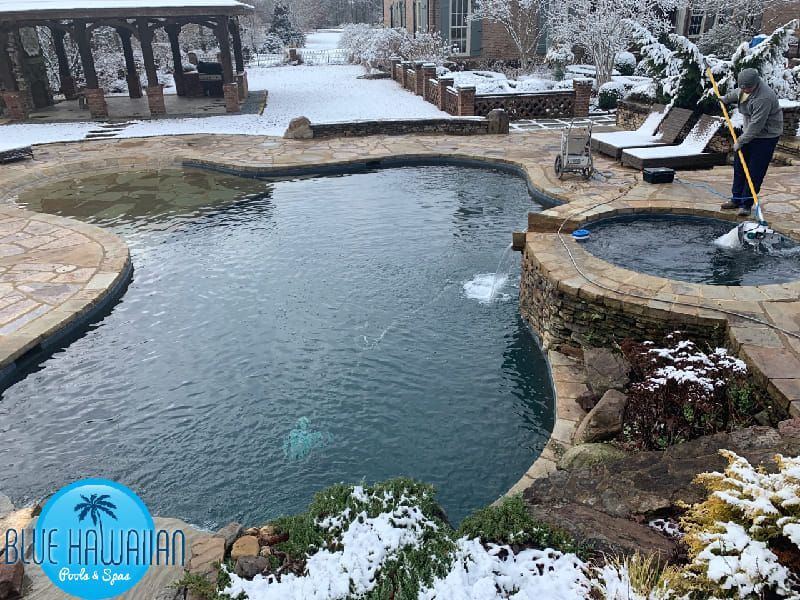 A person cleaning a pool in the snow, a gazebo and spa, surrounded by stone and winter landscaping.