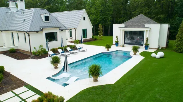 White house with pool and lounge chairs, green grass.