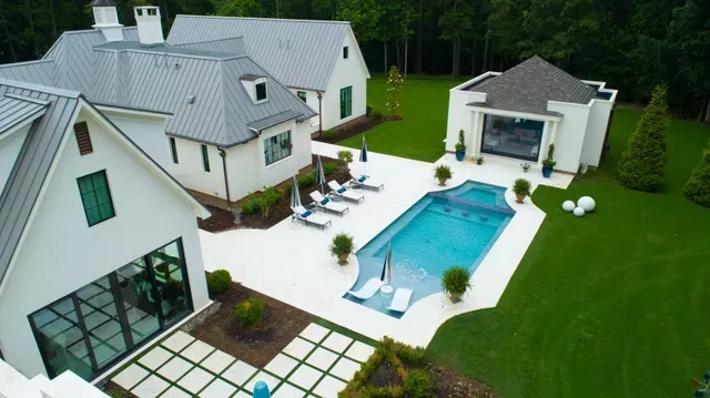 Aerial view of a luxury home with pool, sun loungers, and lush green lawn. White exterior, blue pool.