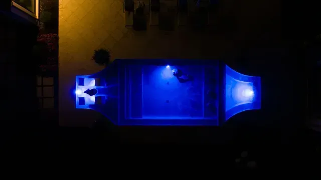 Overhead view of a blue-lit swimming pool at night, with lights in the corners and a figure in the water.