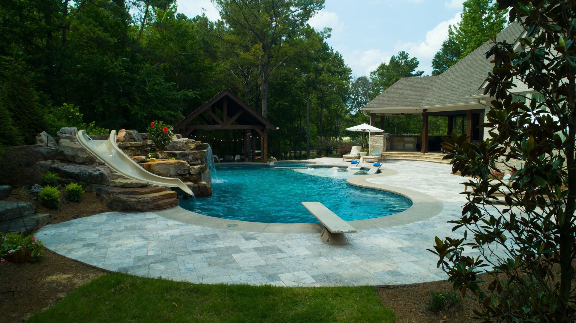Backyard pool with a slide, diving board, and patio area next to a house and wooded area.