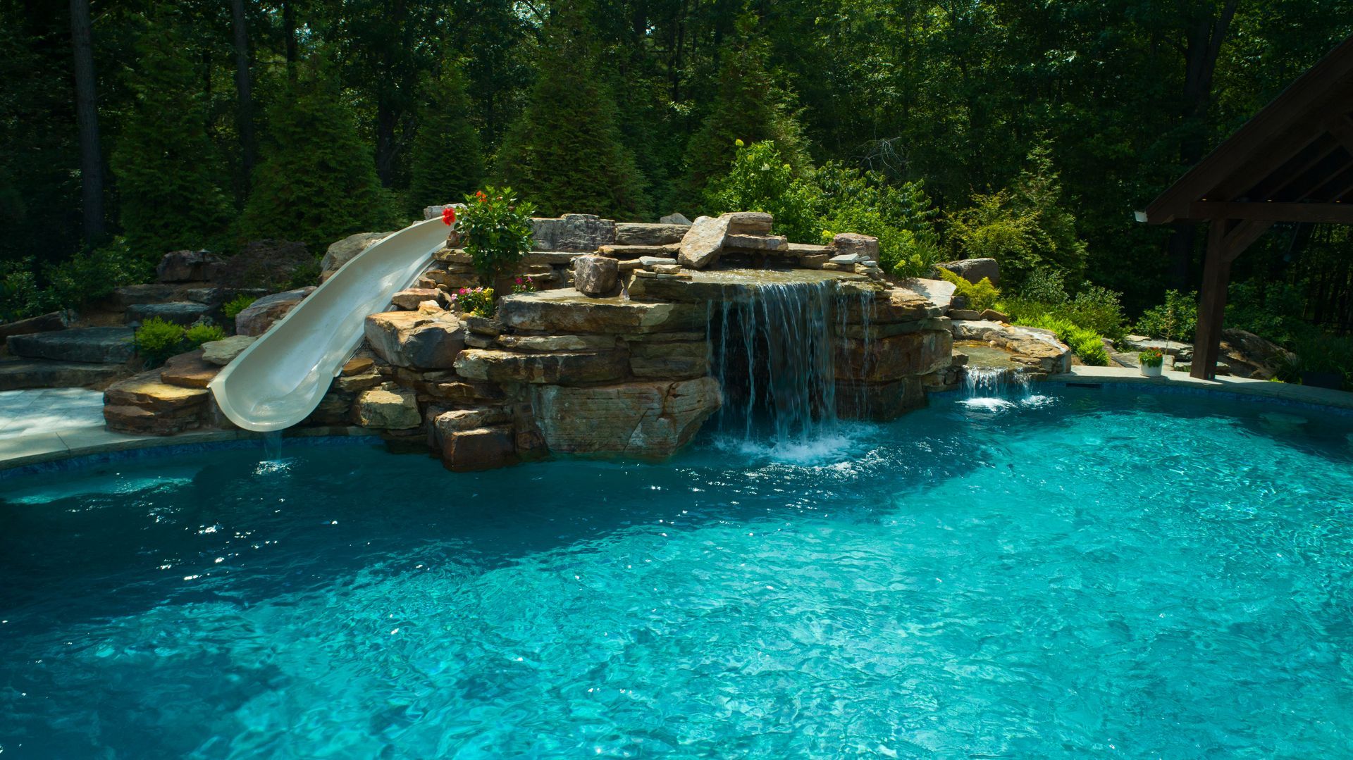 Swimming pool with a rock waterfall and slide. Blue water, green trees in the background.