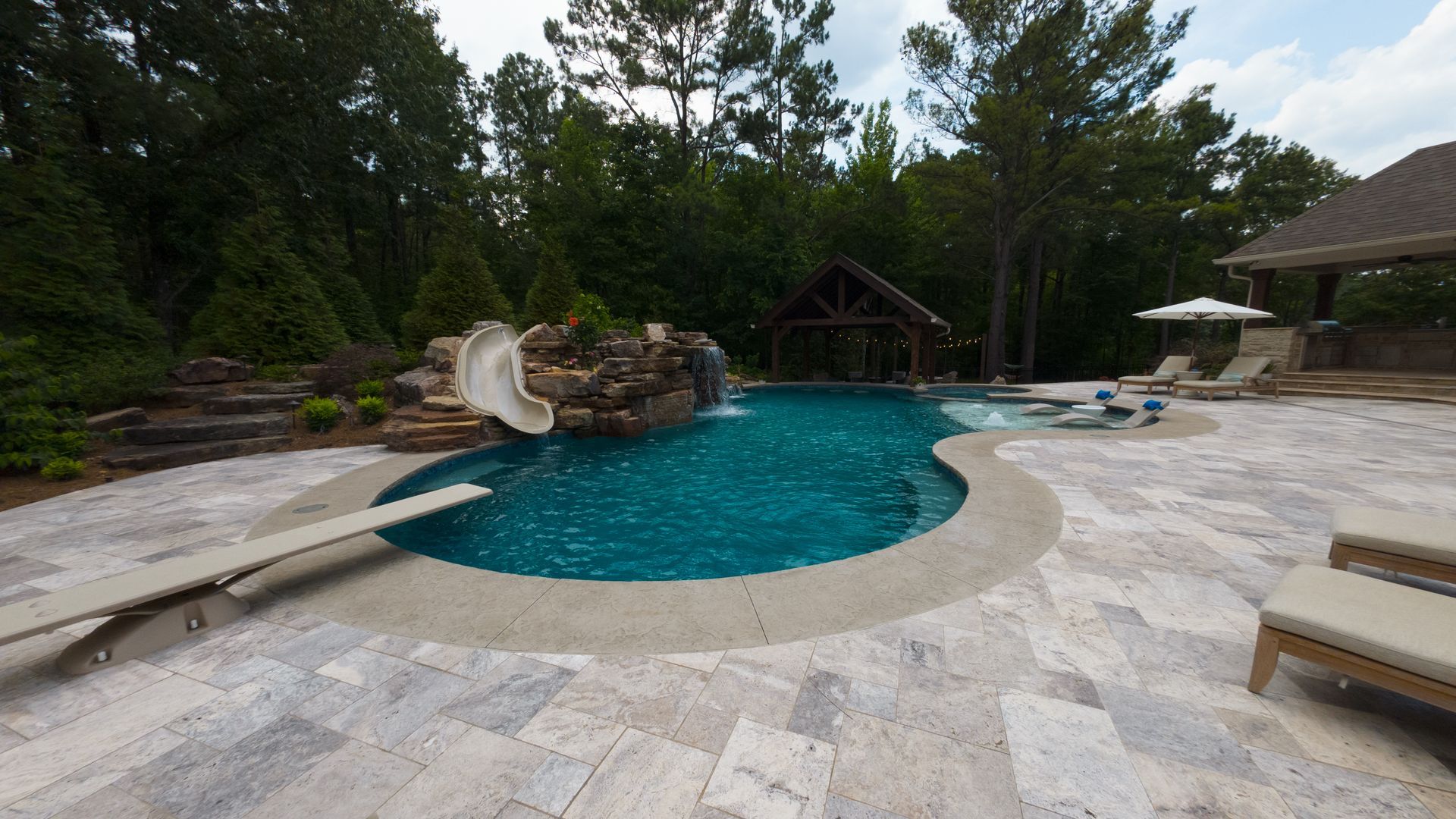 A luxurious backyard pool with a slide, diving board, and gazebo, surrounded by trees.