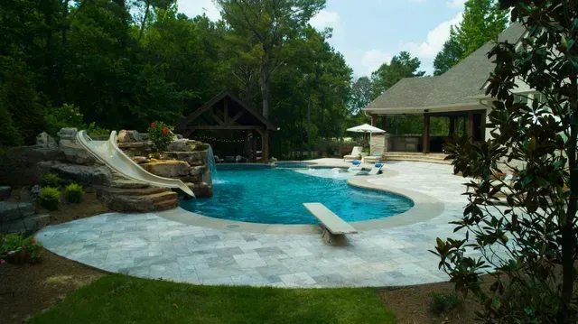 Pool with rock slide, diving board, and covered patio in a backyard setting with trees.