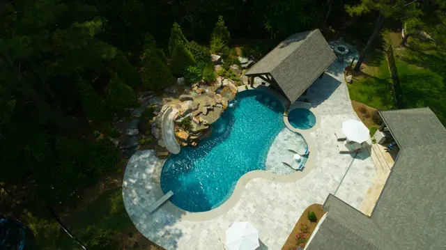 Overhead view of a luxury pool area with a turquoise pool, hot tub, and stone patio surrounded by greenery.