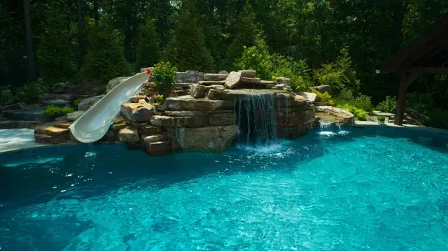 Swimming pool with waterfall feature and slide. Blue water, rock formation, trees in the background.