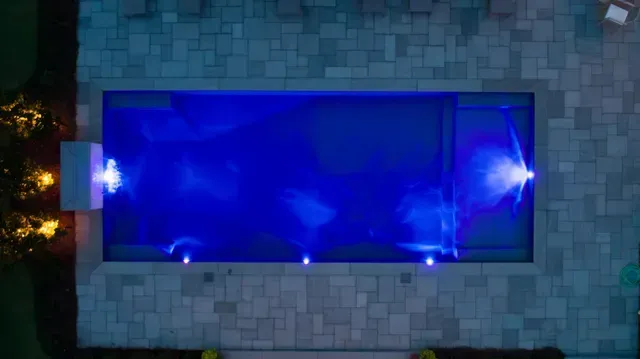 Overhead view of a rectangular pool lit with blue lights at night, surrounded by stone patio.