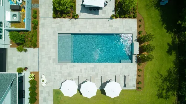 Aerial view of a rectangular pool with sun umbrellas, stone patio, and green lawn.